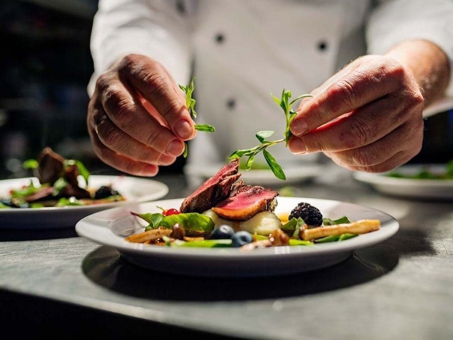 A chef garnishing plates of seared meat and vegetables